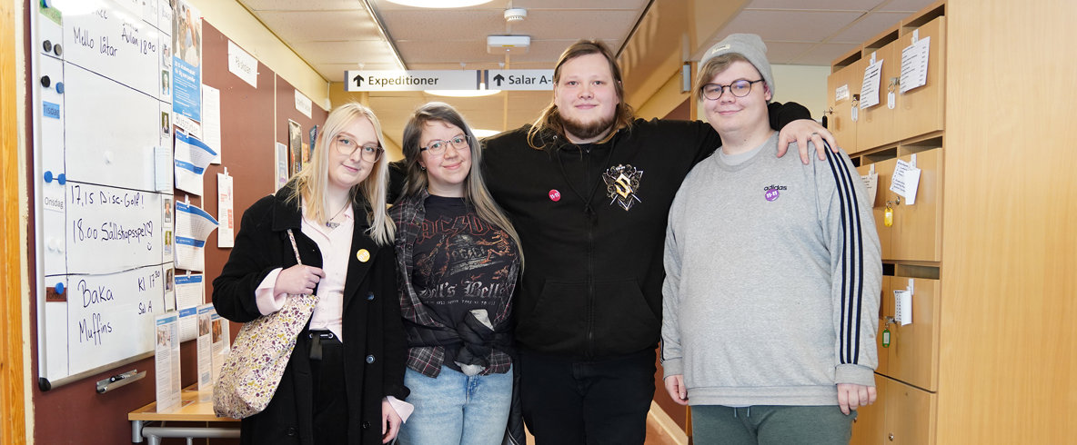 Fyra glada ungdomar på rad håller om varandra i skolkorridor.